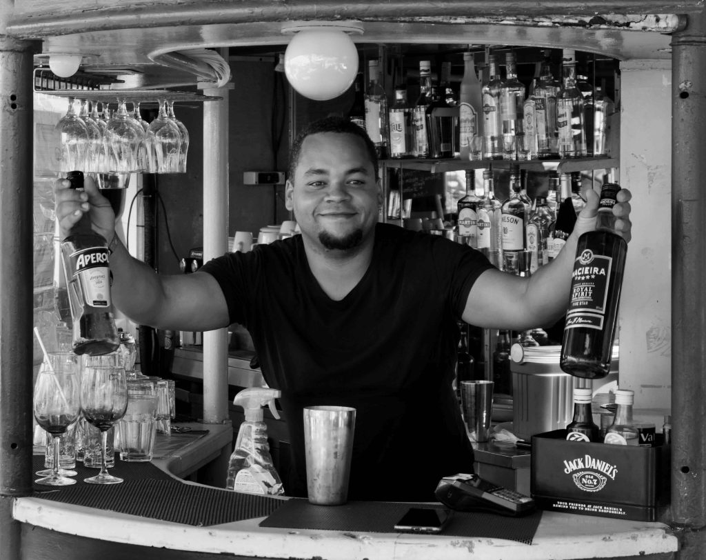 Black-and-white photograph of a smiling bartender behind a bar holding two bottles, with glasses and spirits on shelves behind him.