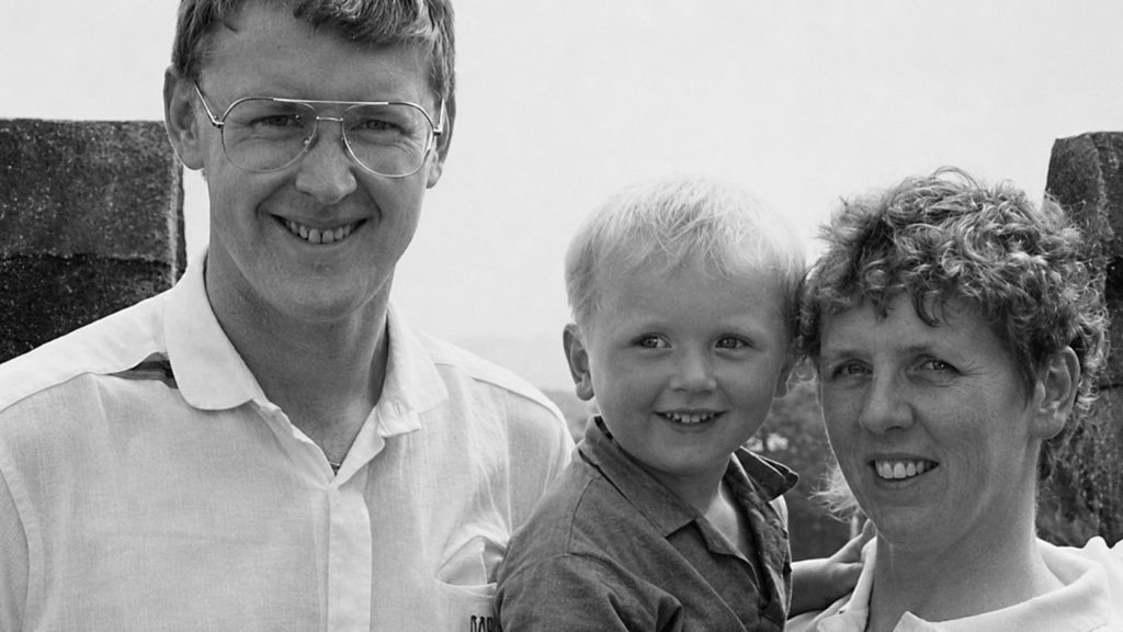 Black-and-white close-up portrait of Sue and Tony standing with a young Scott between them, all looking towards the camera outdoors