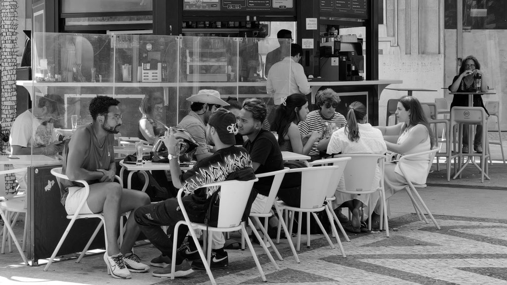Black-and-white photograph of people sitting at outdoor café tables beside a busy coffee kiosk.