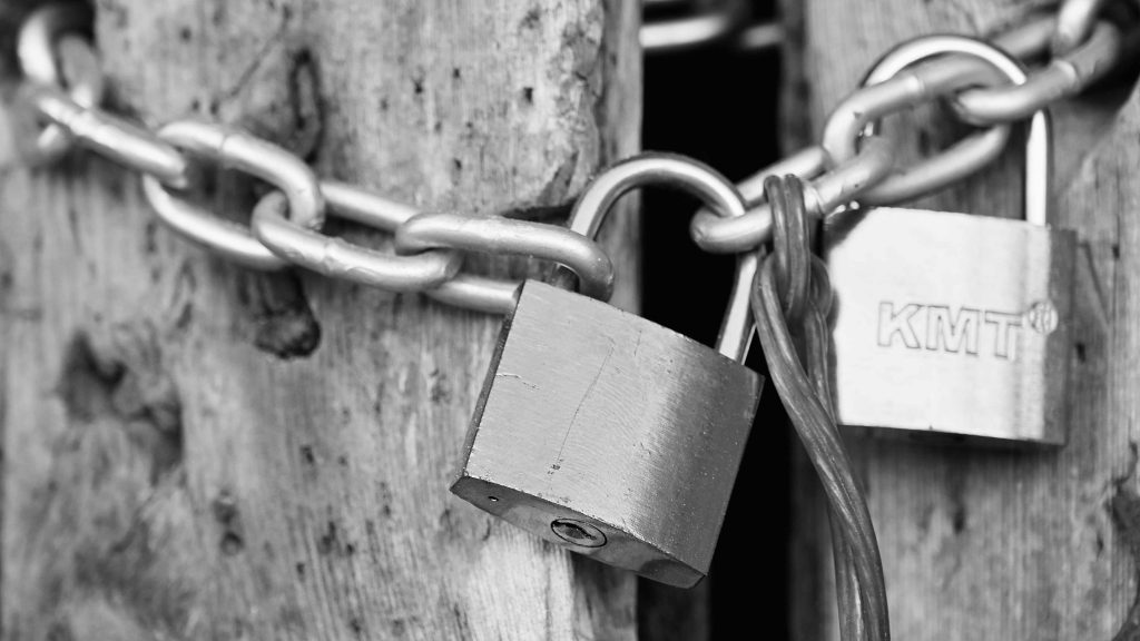 Close-up black-and-white photograph of metal padlocks and a chain securing weathered wooden doors.
