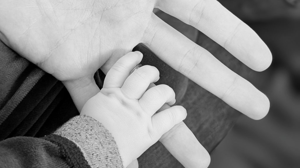 A baby’s small hand resting in an adult’s open palm, photographed in close-up in black and white.